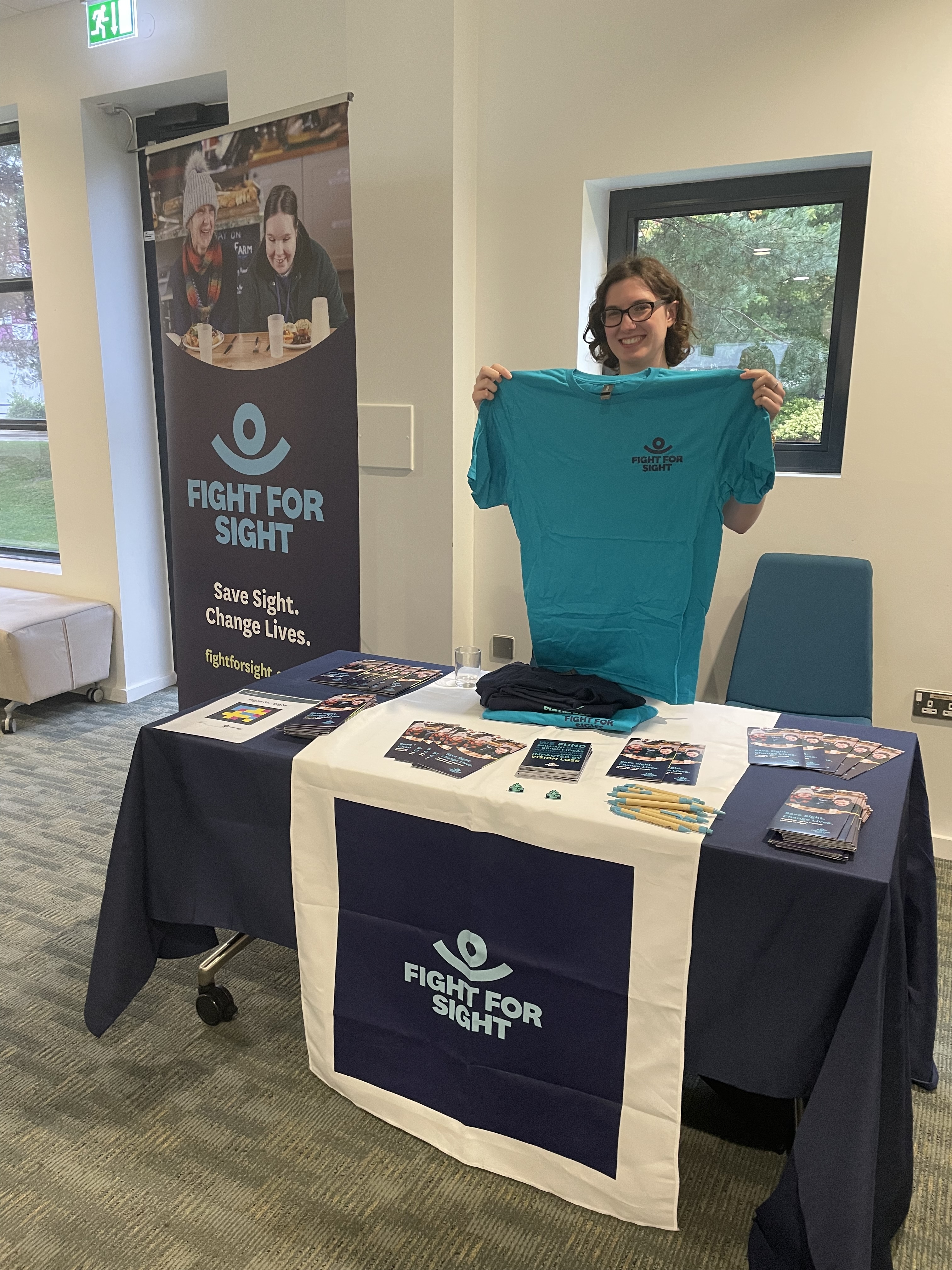Eleanor Durrant stands behind the Fight for Sight stand at Visionary 2024 surrounded by new merchandising and holding up a branded tshirt