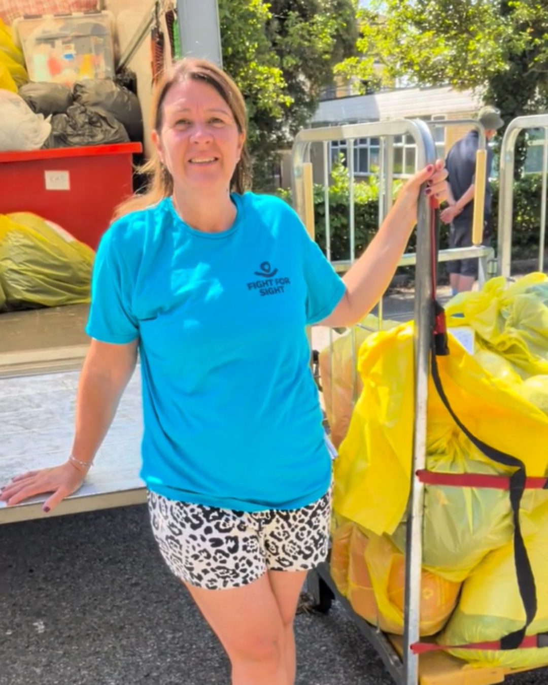 A photo of Emma Salisbury from the Tommy Salisbury Choroideremia Family Fund. Emma wears a blue fight for sight tshirt and stands in front of a van full of items for donation, she is holding a tall trolley full of yellow bags full of even more donations.