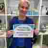 Christine Hampton Hill charity shop volunteer, she has beautifully styled silver hair and a stylish blue jacket and is holding up a sign that says Yes please Hampton Hill! We'd love your donations!