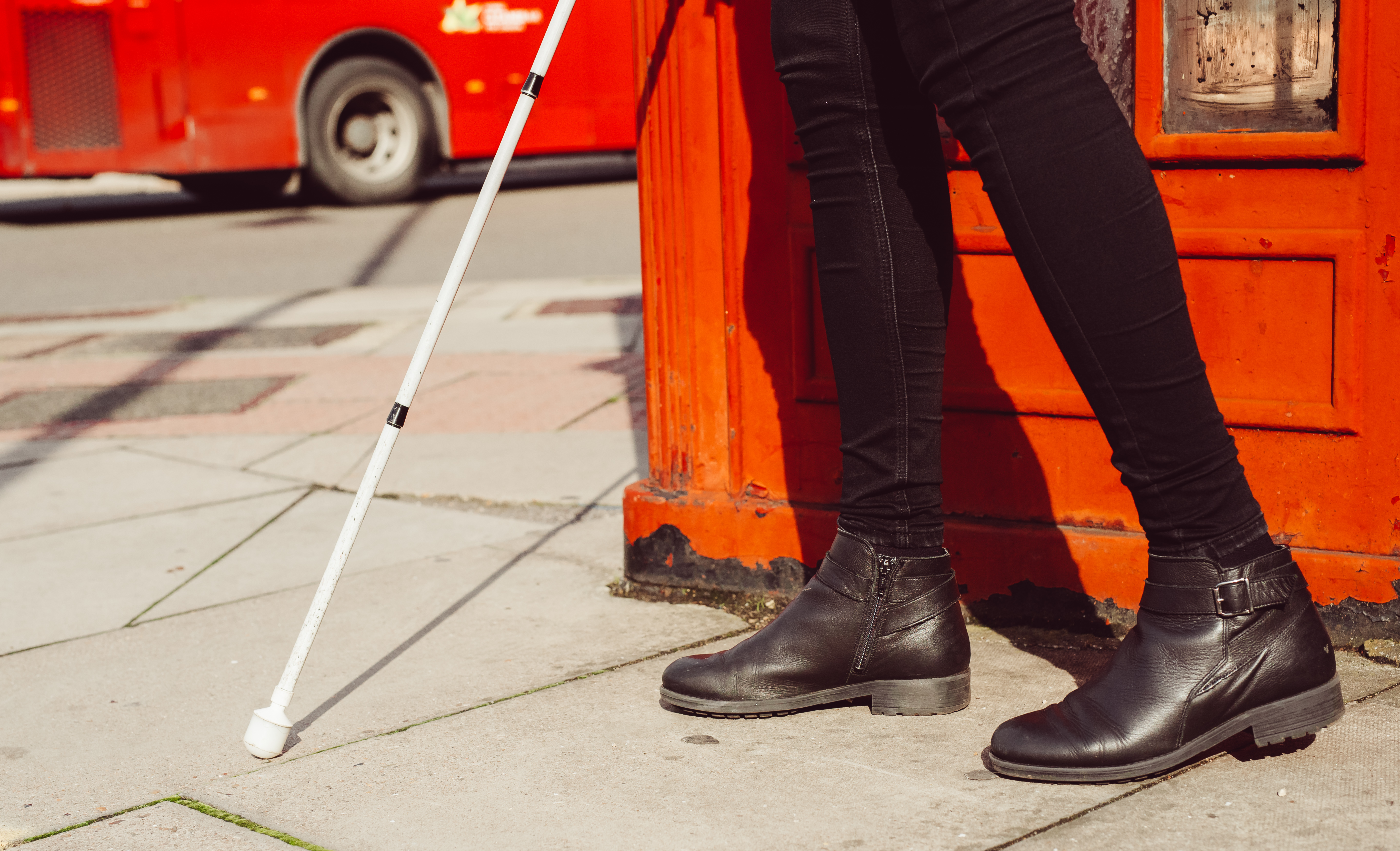 A shot from the knees down of someone using a white cane. A telephone box and red bus can be seen in the background.