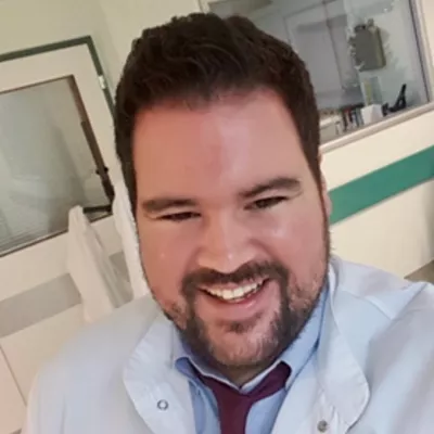 George is smiling open mouthed at the camera. He's wearing a tie and white lab coat. 