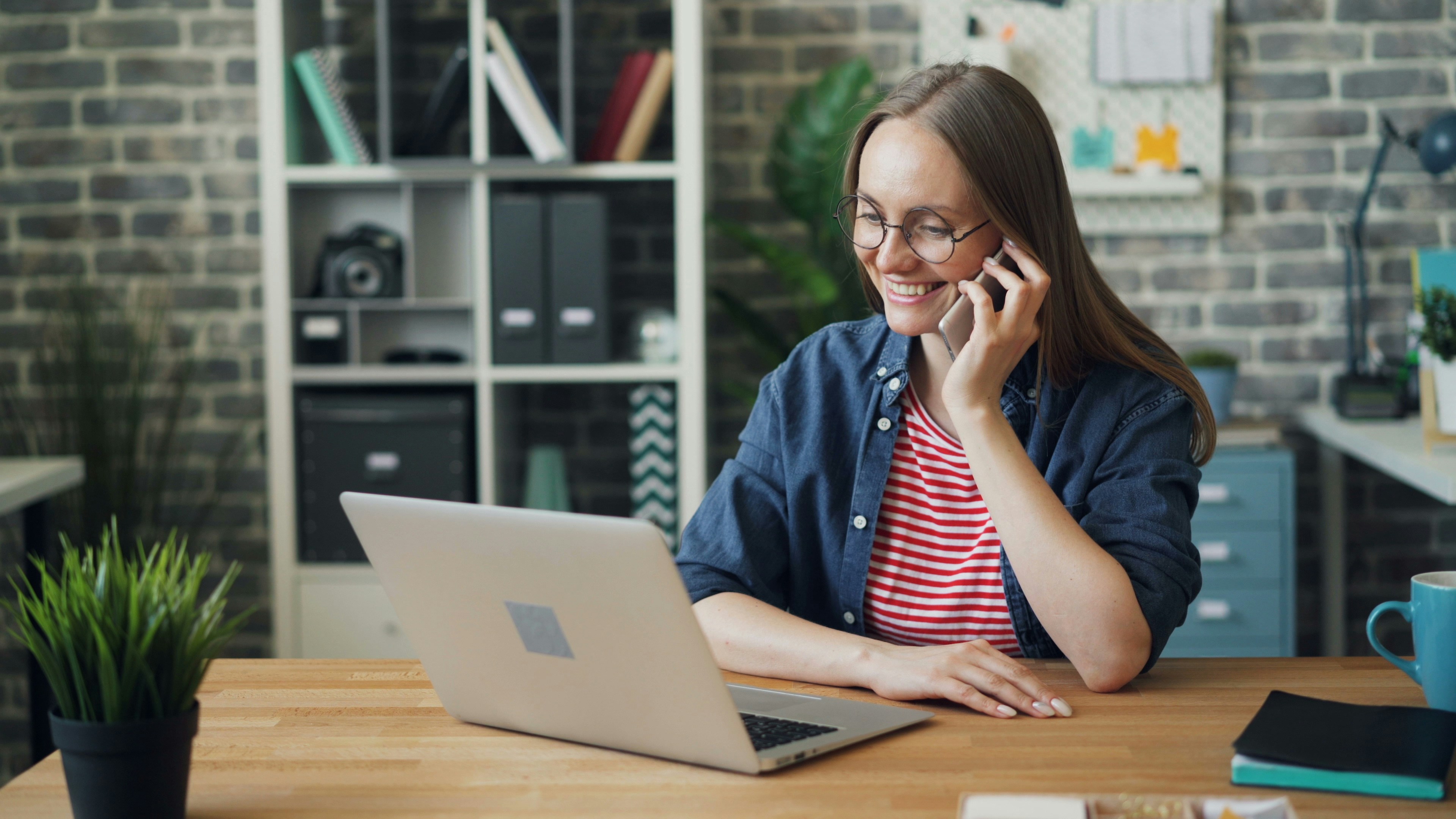 A woman on the phone, she is smiling and looking down at a laptop screen