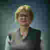 Mary-Anne is a white woman with blonde hair and glasses. She wears a green tank top over a shirt. 
