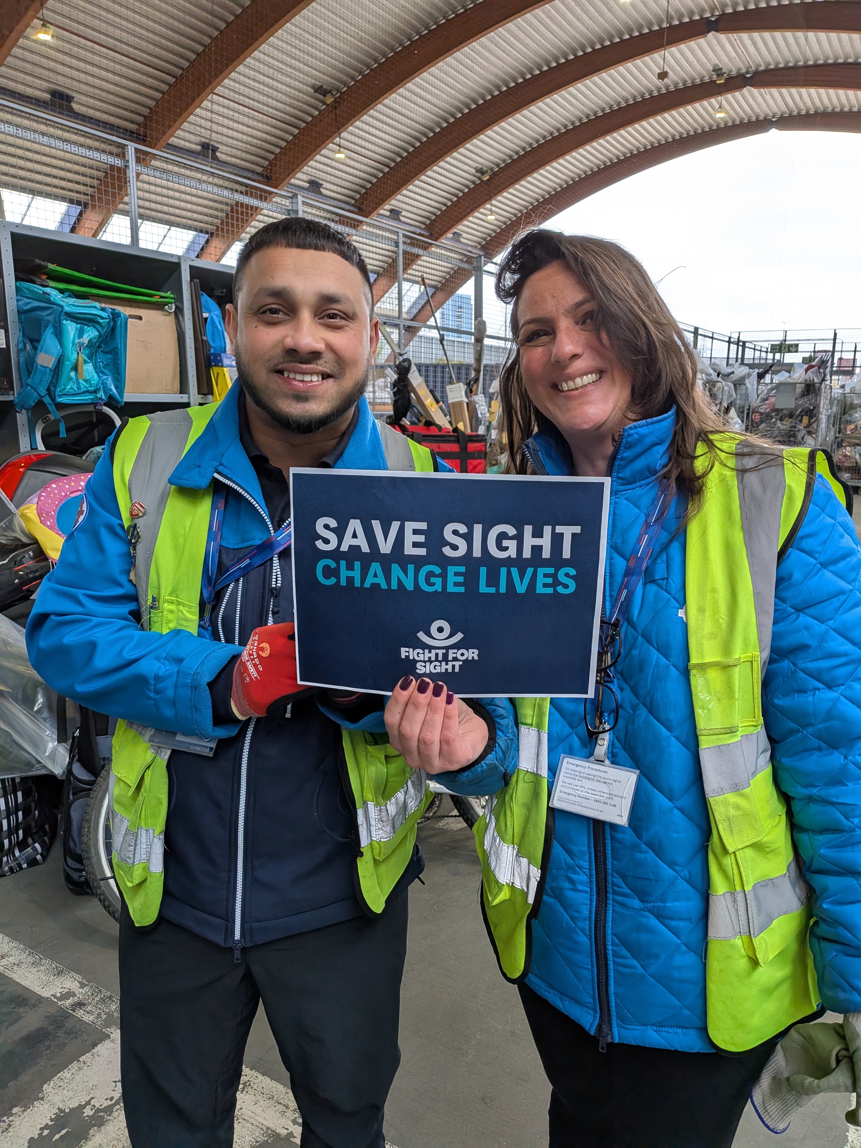 TfL Lost Property donation to Fight for Sight man and lady holding up a sign at the Lost Property Depot that says Save Sight. Change Lives.