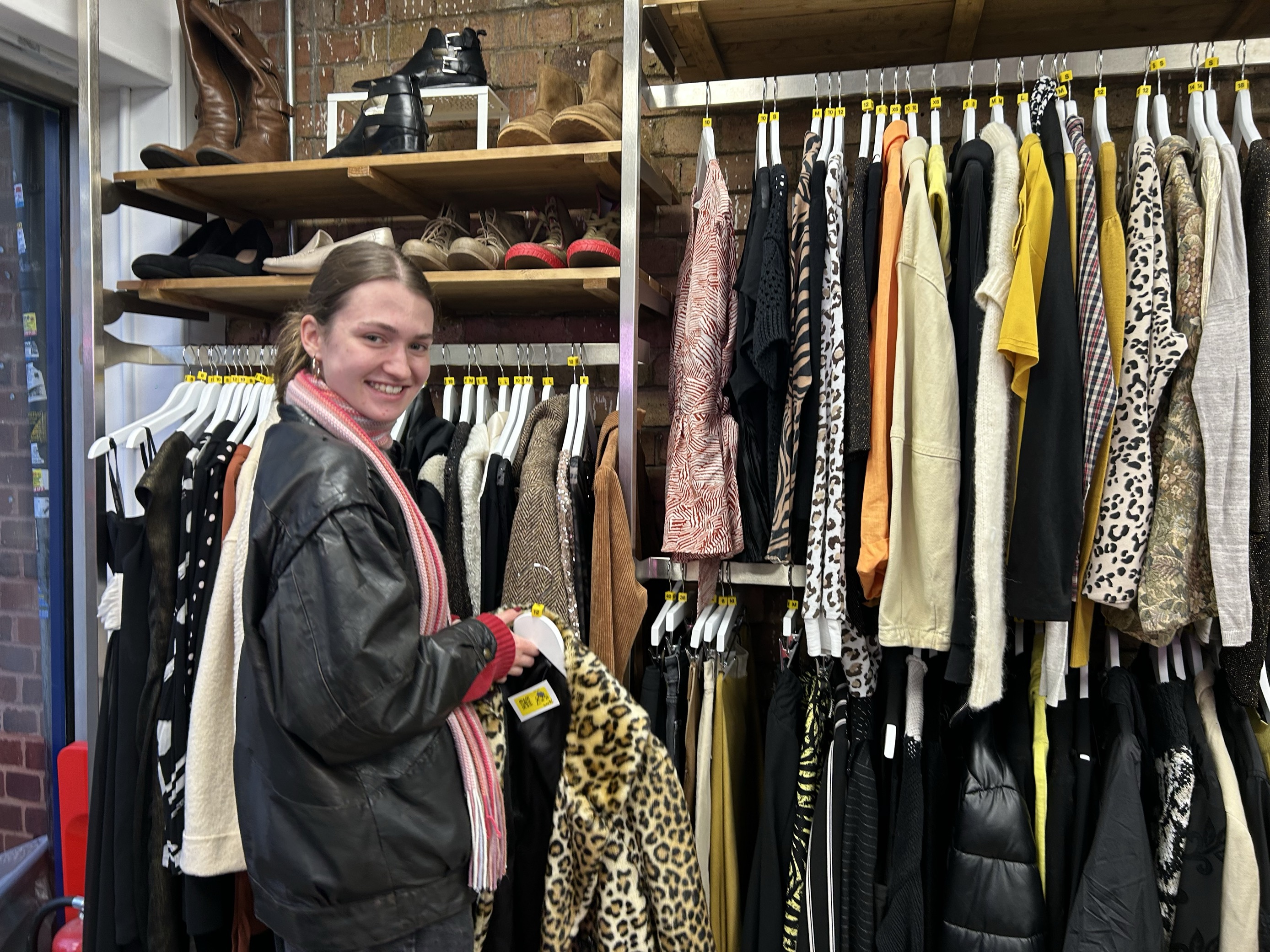 Evie is a young woman in a leather jacket and a pink scarf, smiling as she holds up a leopard print fur coat.