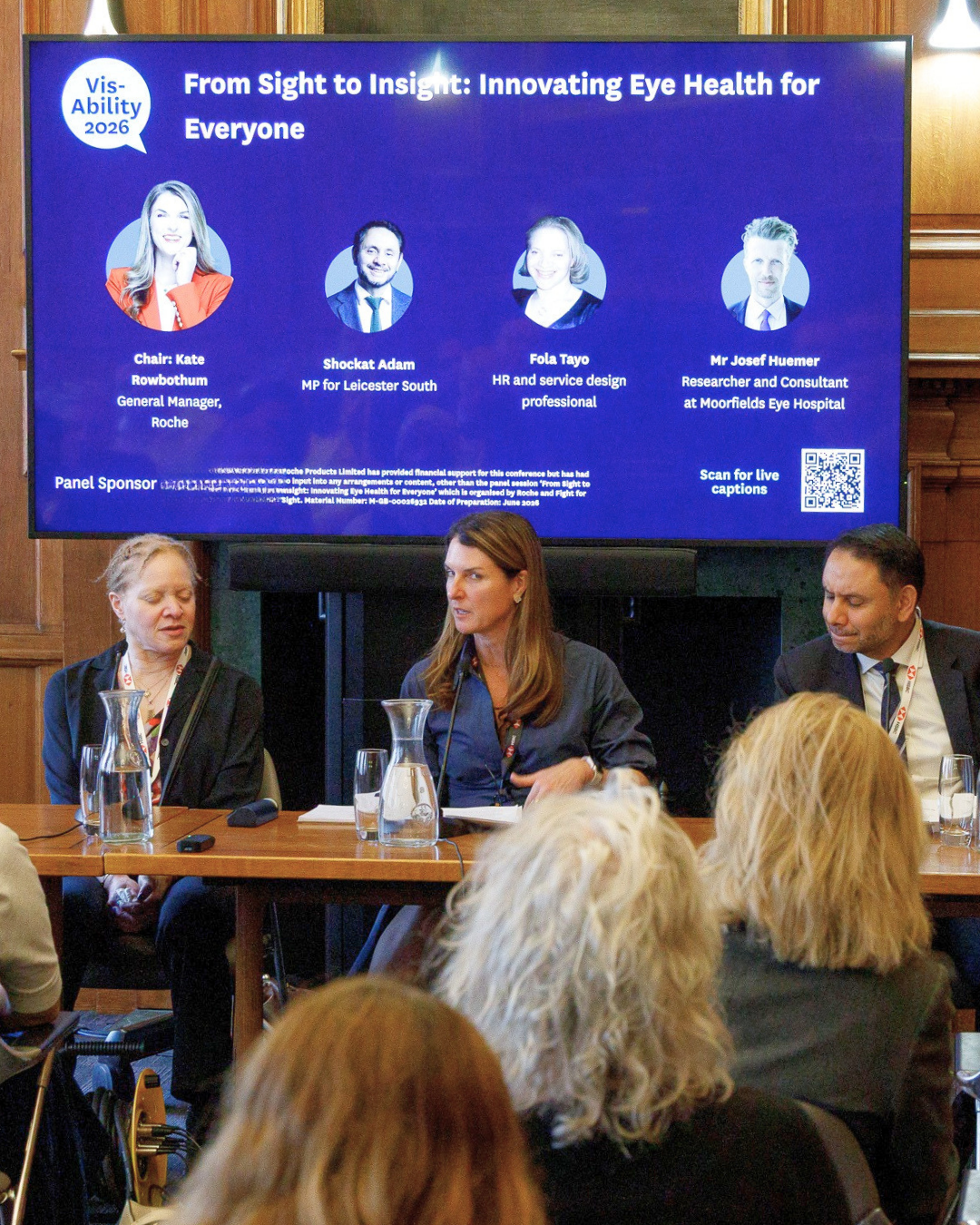 VisAbility innovation in sight loss research session where three people are shown holding a discussion in front of a large screen in front of a live audience in a wood panelled room