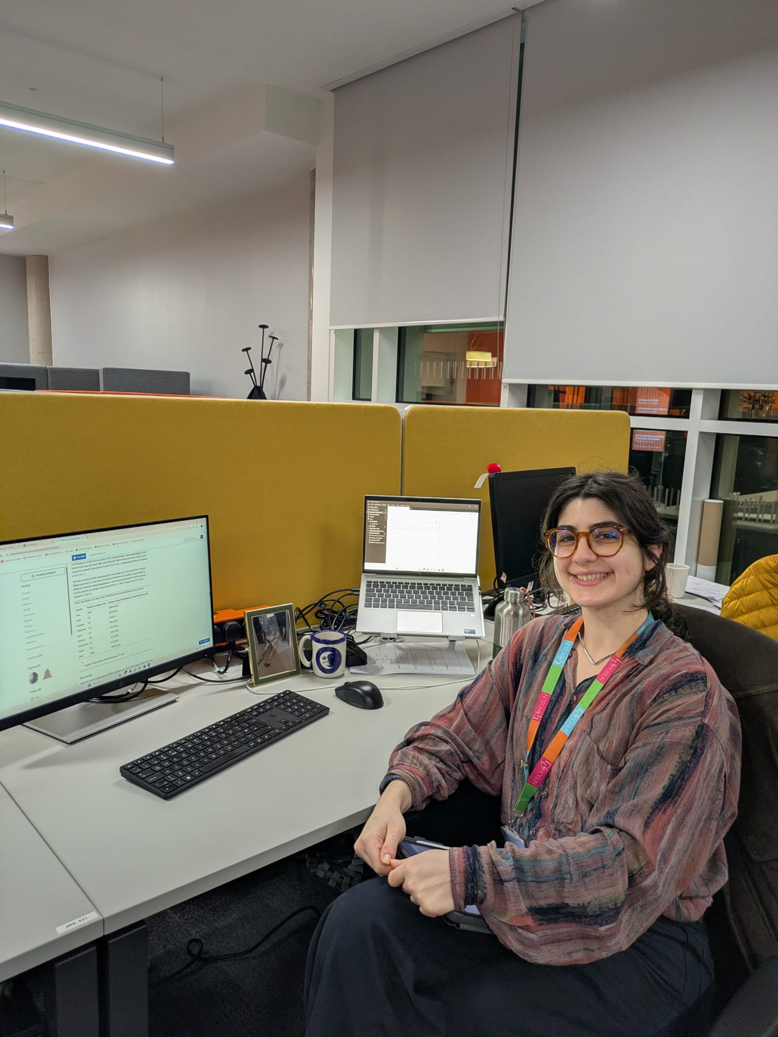 A photo of AJ at their desk at university. AJ is wearing tortoise shell glasses, a loose fitting shirt with their hands holding a tablet in their hand. 