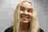 Kerry is a woman with shoulder-length blond hair is pictured against a light grey wall. She appears to be looking upward and slightly to her right, with a wide grin that exposes her teeth. Her expression suggests amusement or playfulness. She is wearing a black, possibly knit, cardigan over a black top. A small, delicate necklace is visible. The lighting is somewhat diffused, casting soft shadows and drawing attention to her face. The wall's texture is slightly rough.