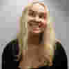 Kerry is a woman with shoulder-length blond hair is pictured against a light grey wall. She appears to be looking upward and slightly to her right, with a wide grin that exposes her teeth. Her expression suggests amusement or playfulness. She is wearing a black, possibly knit, cardigan over a black top. A small, delicate necklace is visible. The lighting is somewhat diffused, casting soft shadows and drawing attention to her face. The wall's texture is slightly rough.