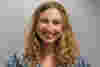 Nina has light brown curly hair and a smile is pictured against a light grey background, wearing a blue and white checked shirt.