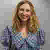 Nina has light brown curly hair and a smile is pictured against a light grey background, wearing a blue and white checked shirt.