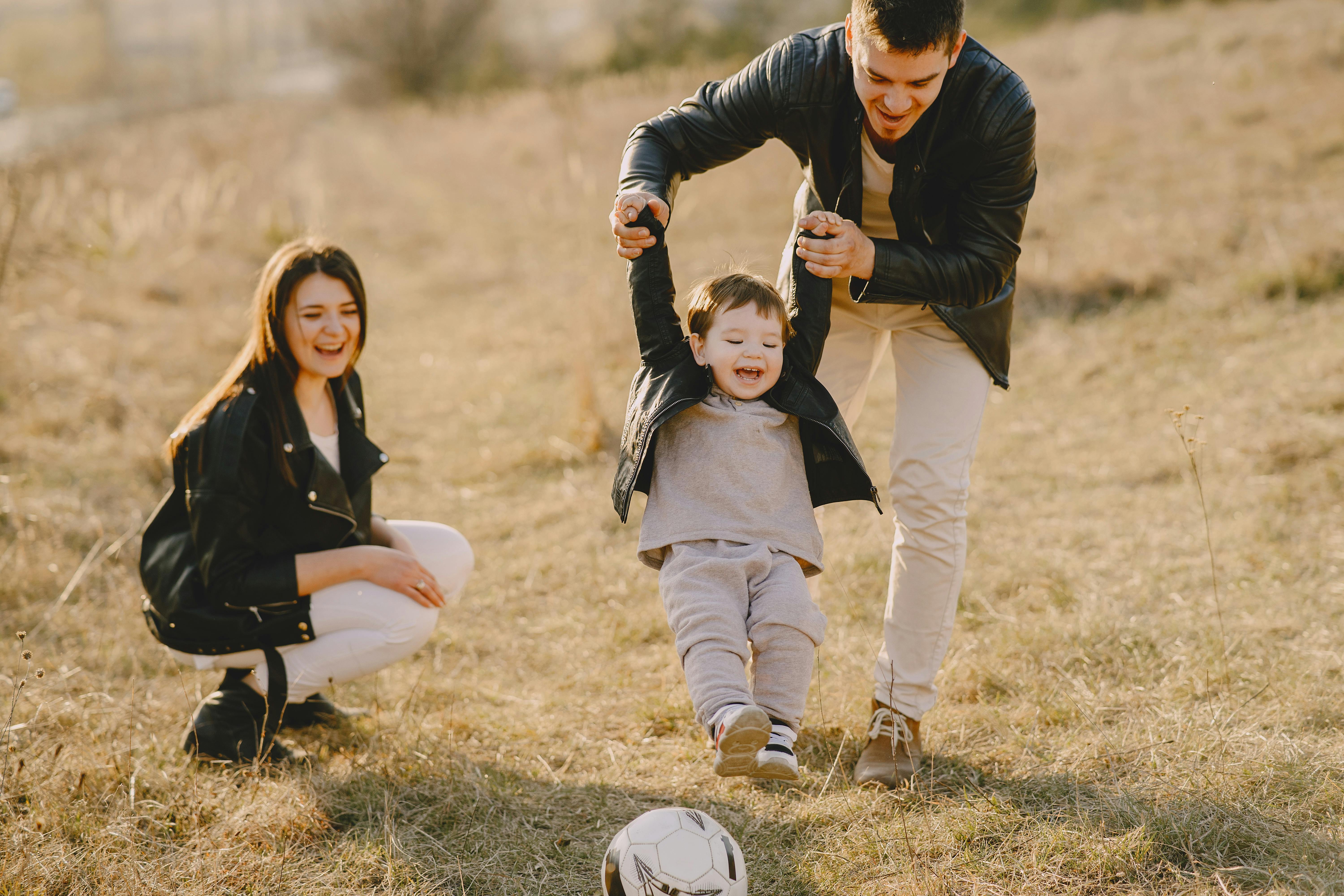 A mum and dad in a field with their child who is trying to kick a football