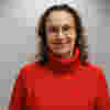 Jo has shoulder-length, wavy brown hair is smiling and wearing glasses. She is wearing a red turtleneck sweater. The background is a light gray wall. The light source is slightly above and to the front of the woman. The texture of the sweater is noticeable.  
