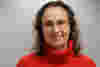 Jo has shoulder-length, wavy brown hair is smiling and wearing glasses. She is wearing a red turtleneck sweater. The background is a light gray wall. The light source is slightly above and to the front of the woman. The texture of the sweater is noticeable.  