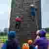 A group of children wearing bright helmet scale an outdoor climbing wall