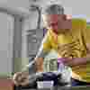 Clarke in his studio, creating a piece for one of his upcoming shows. It's a bright room with artwork in the background. Clarke is wearing a yellow tshirt and concentrating hard on his work