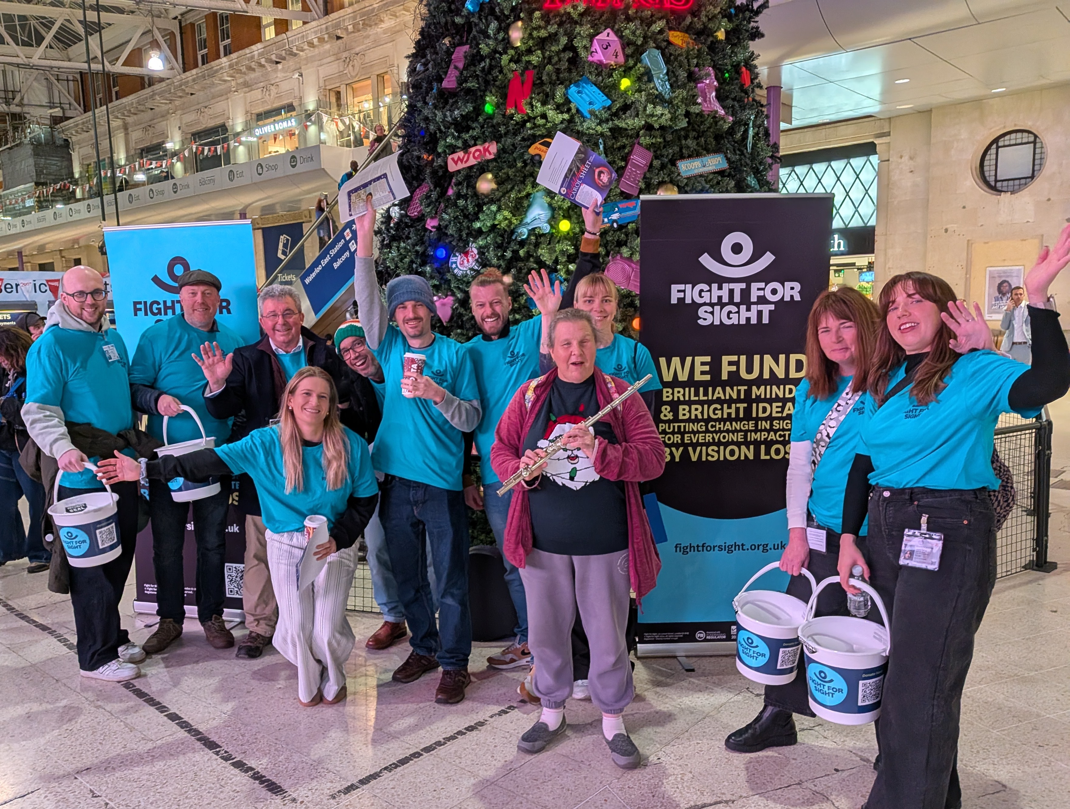 Fight for Sight Christmas Carol Singers and volunteers at Waterloo Station standing in front of a huge Christmas tree