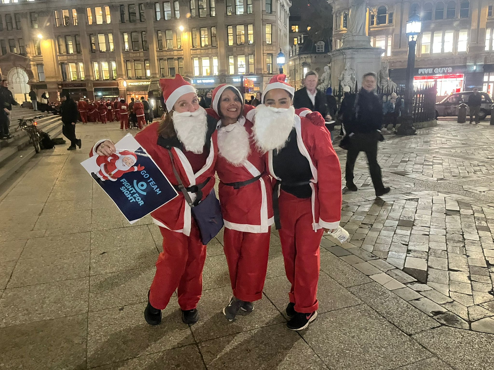 Fight for Sight supporters wearing a Santa suit and proudly displaying a Fight for sight poster, as part of Santa in the City.