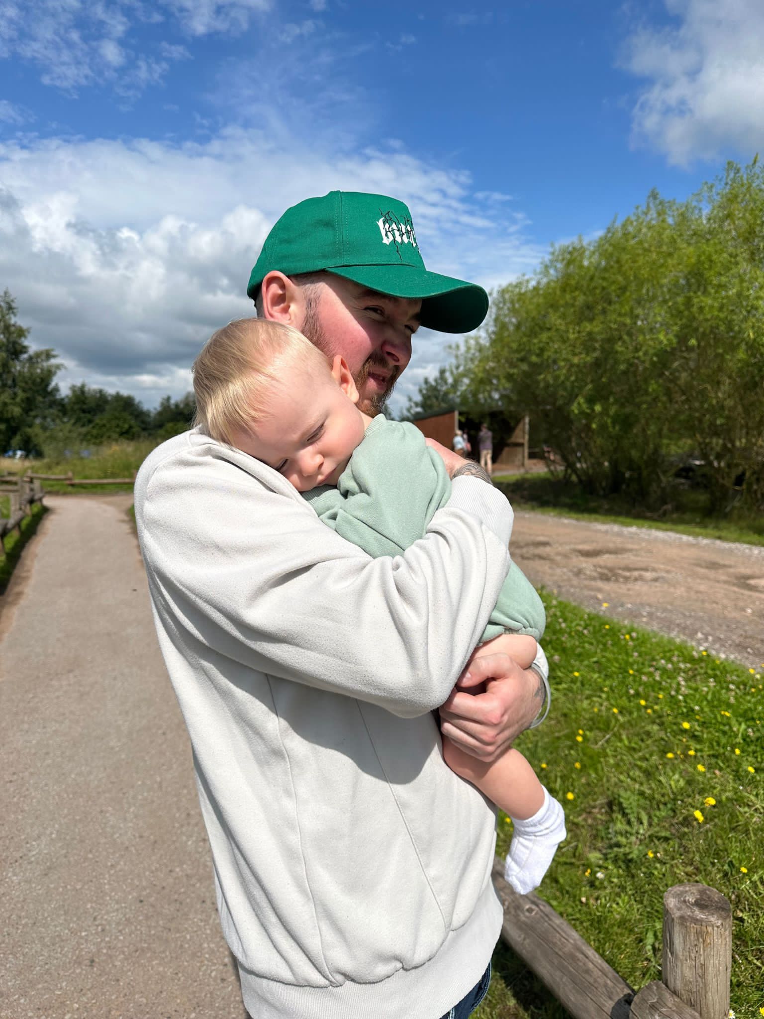 Greg, a white man in his late 20s with a dark beard and wearing a green baseball caps is standing on path on the edge of a park. He's cuddling his baby nephew who is asleep. The baby is wearing a green vest with white socks and looking very content and snug.
