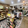 A well-organised retail store with a mix of homeware, clothing, and accessories. The store features wooden shelving filled with glassware, ceramics, and framed artwork. A central display table holds decorative candles and colourful textile items. In the background, clothing racks showcase a variety of garments, while industrial pendant lights hang from the ceiling, adding to the store’s modern aesthetic.