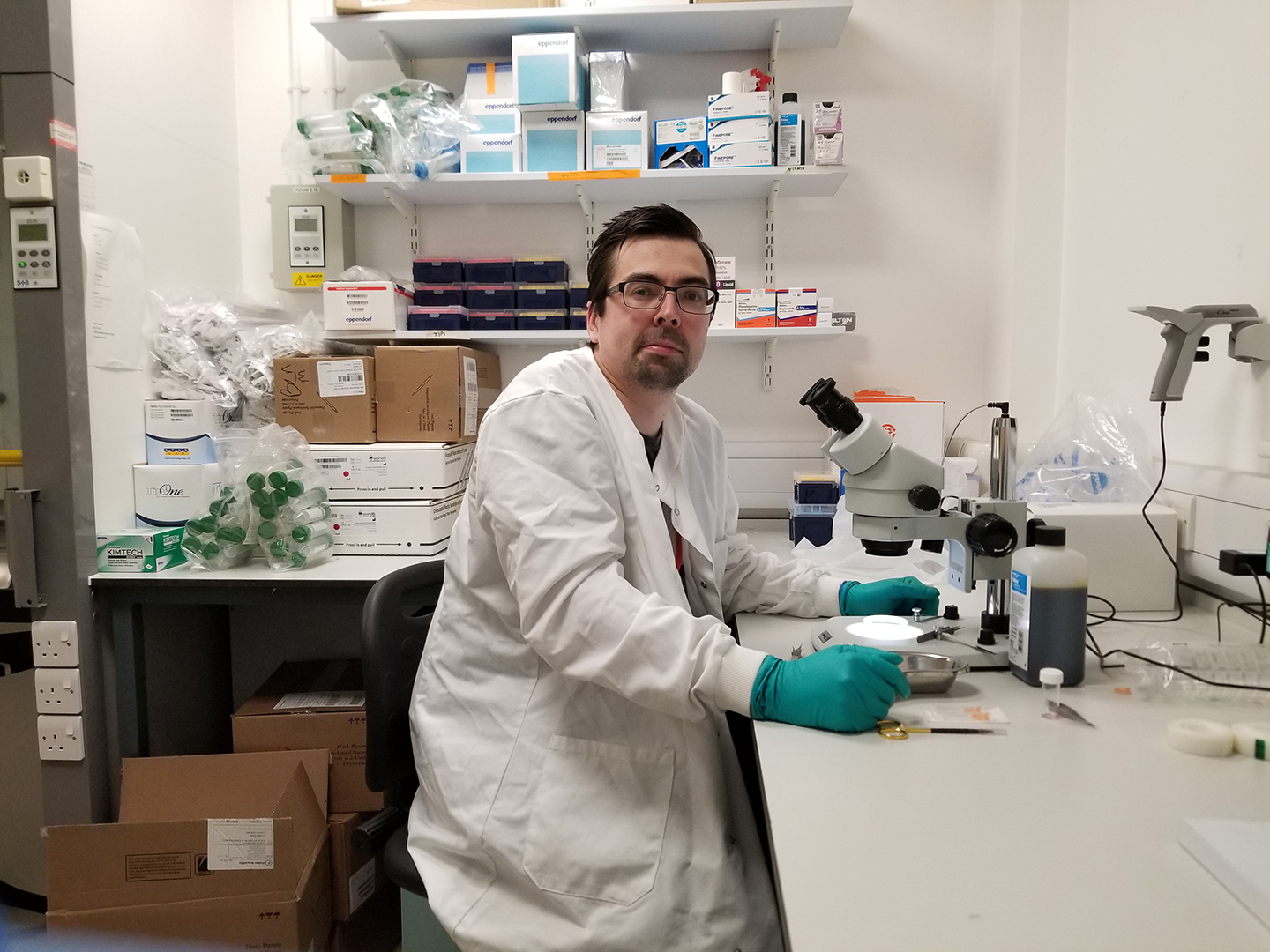 Dr Ben Mead lecturer and researcher at Cardiff University’s School of Optometry and Vision Sciences in a lab sitting at a microscope wearing a white lab coat.
