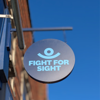 Fight for Sight hanging sign, a navy blue circle with our logo in the centre, with a blue sky behind. 