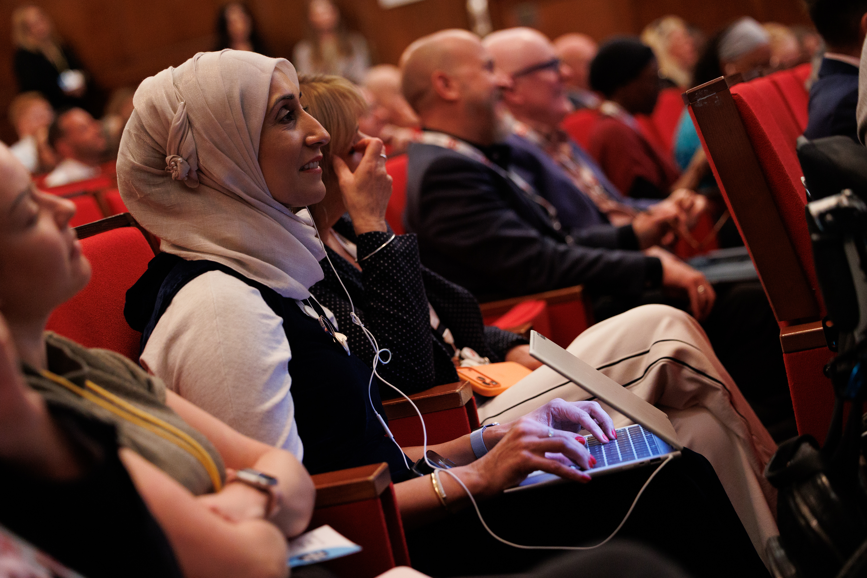 A woman in a hijab looks ahead while typing into a laptop taking notes at an event. In the background are other people listening to what is being said ahead and making notes
