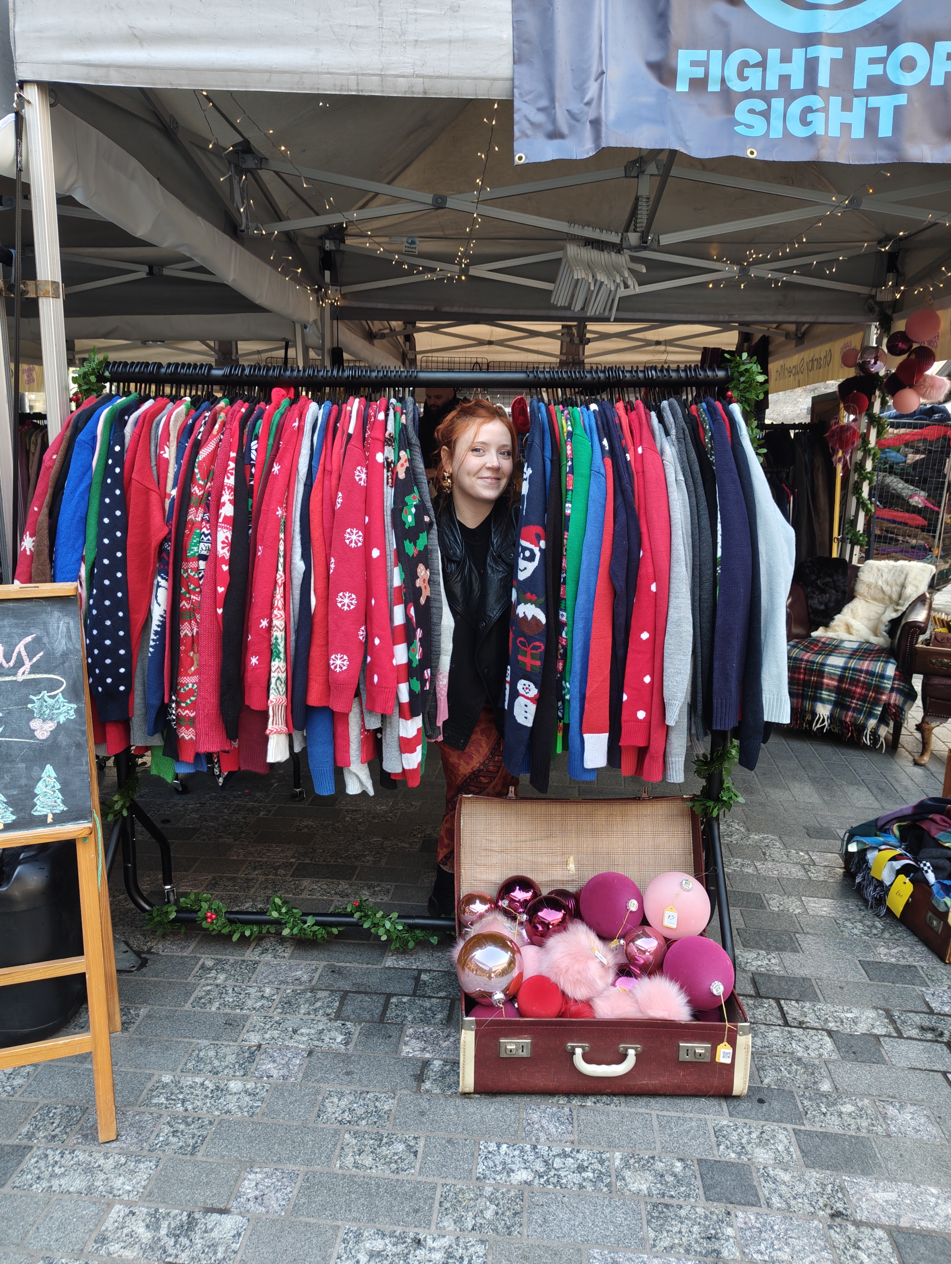 Lauren at the Fight for Sight Coal Drops Yard Christmas Popup - her smiling face peeping out from a rail of Christmas jumpers