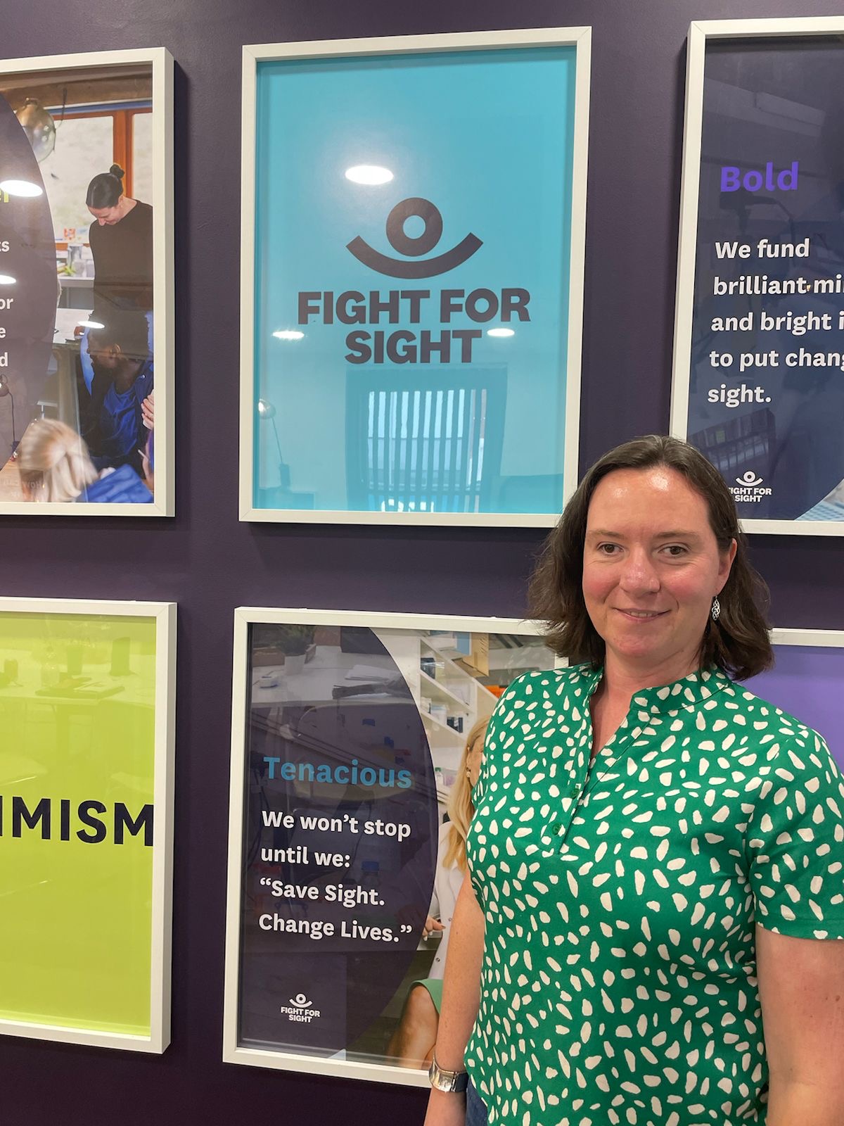 Ellie is standing in front of some posters in our Eaing shop - she is wearing a spotty top and smiling. 