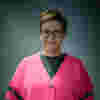 Beck is a white woman with short hair and glasses. She wears a bright pink cardigan. 
