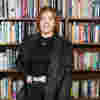 Tilly 'The Blind Stylist' from Wanted Wardrobe is standing in front of a book shelf wearing a black outfit with a sparkly blazer and belt.