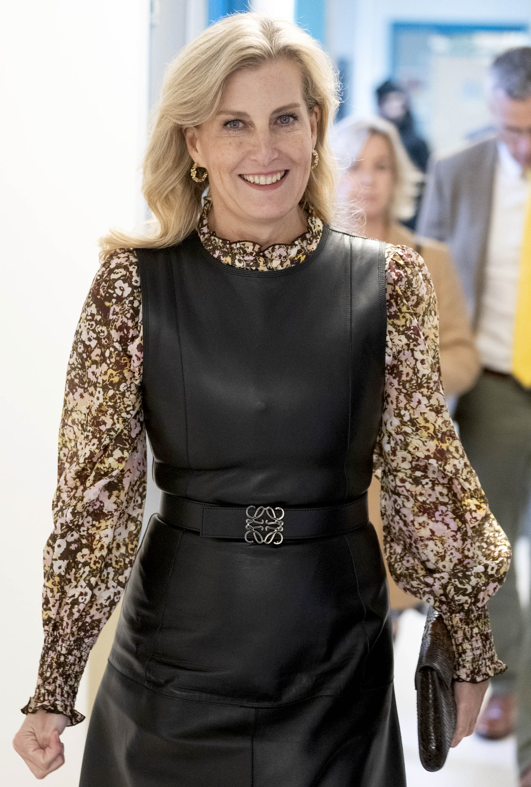 HRH The Duchess of Edinburgh, a blond woman in her late 50s is walking with confidence down a passageway. She's smiling at the camera