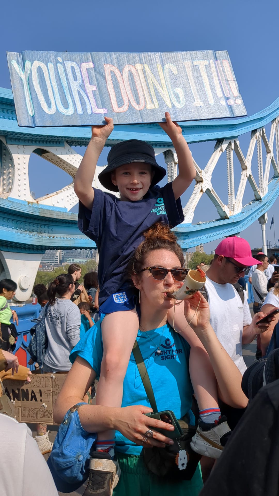 A photo of a boy on a ladys shoulders holding a sign saying 'You're doing it' on Tower bridge during the London Marathon