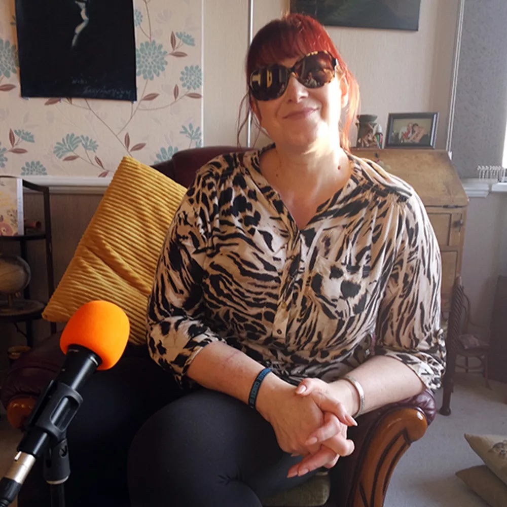 Niki who runs a support group glaucomarize. She is wearing an animal print blouse and sitting behind a yellow microphone. Nike wears dark glasses. 