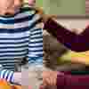 Two women sit at a table holding hands. One woman looks upset while the other woman has her hand on her shoulder and comforting her
