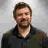 Steven has dark hair and a beard. He wears a dark brown shirt and is smiling for the camera. 
