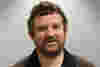 Steven has dark hair and a beard. He wears a dark brown shirt and is smiling for the camera. 