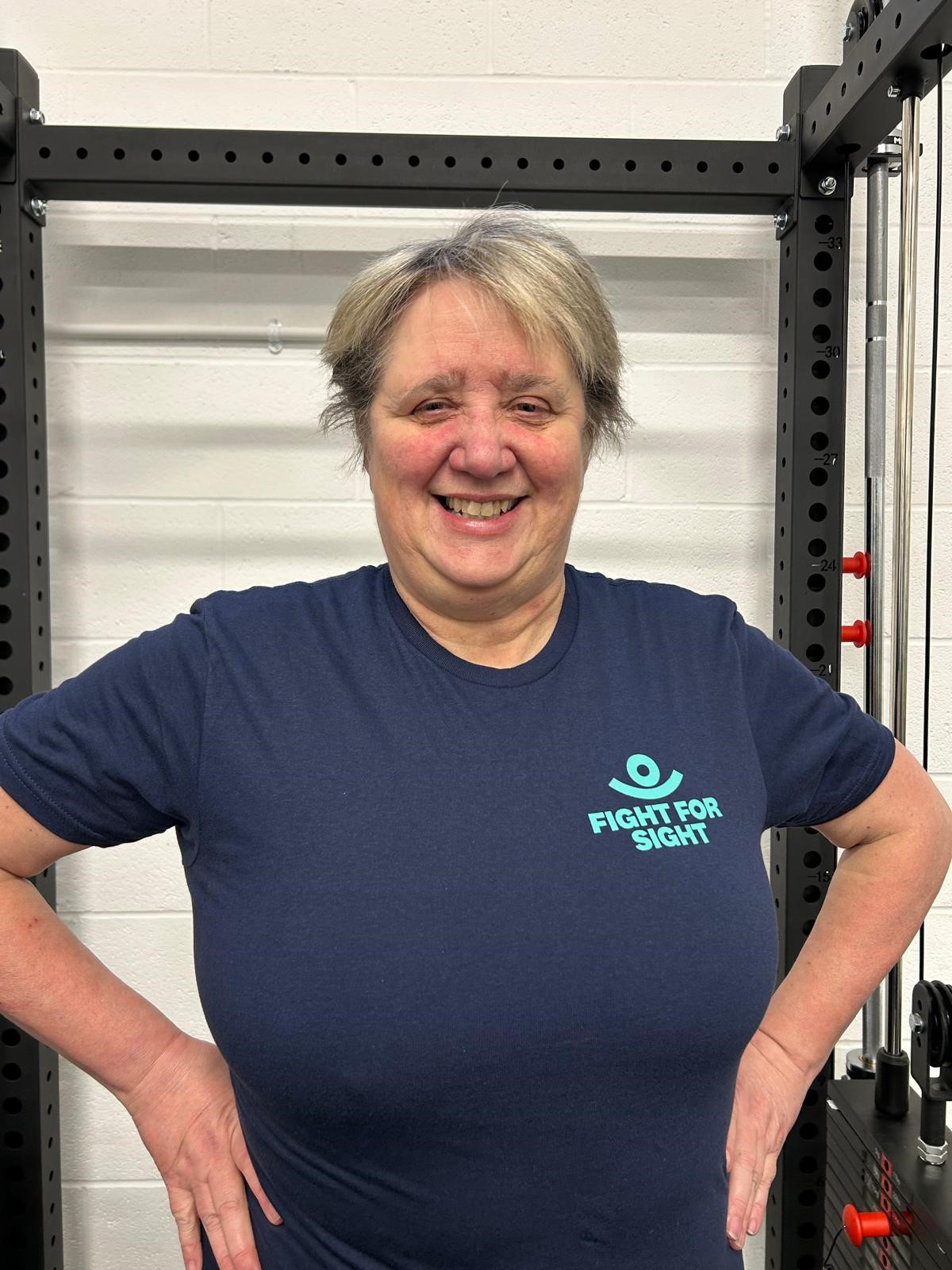 Debbie, a white woman in her 50s, stands proudly in front of gym equipment. She's smiling and wearing a blue Fight for Sight tshirt