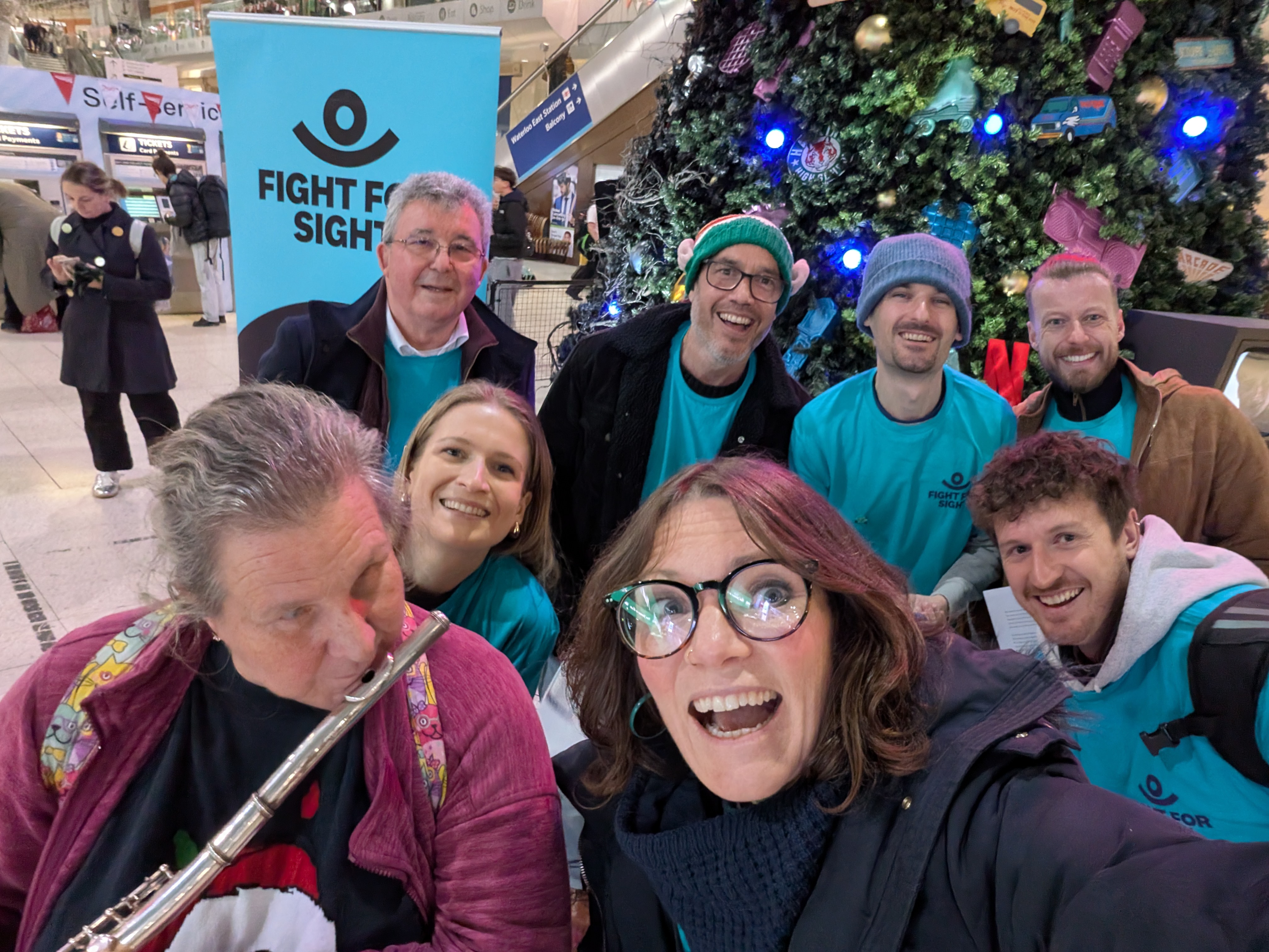 Fight for Sight Christmas Carol Singers at Waterloo Station smiling in front of a huge Christmas Tree