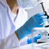 A researcher wearing a coat and mask looks through a microscope