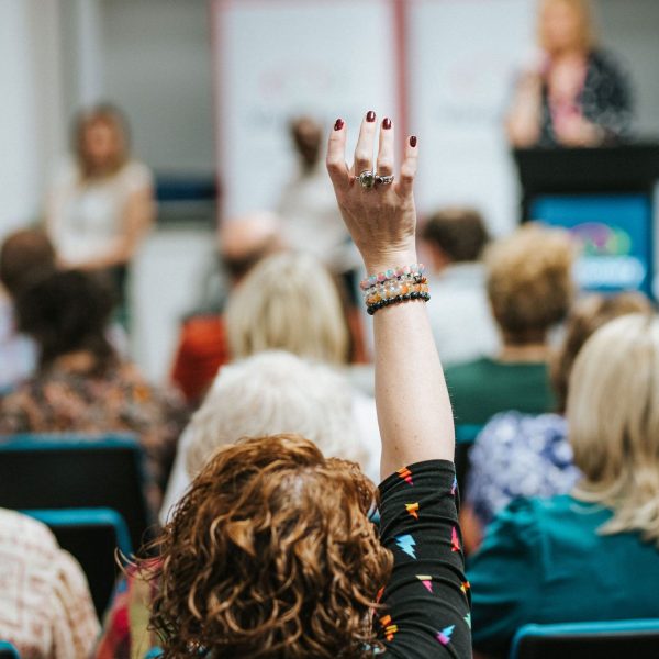 A woman with long red hair sitting in a crowd raises her hand 