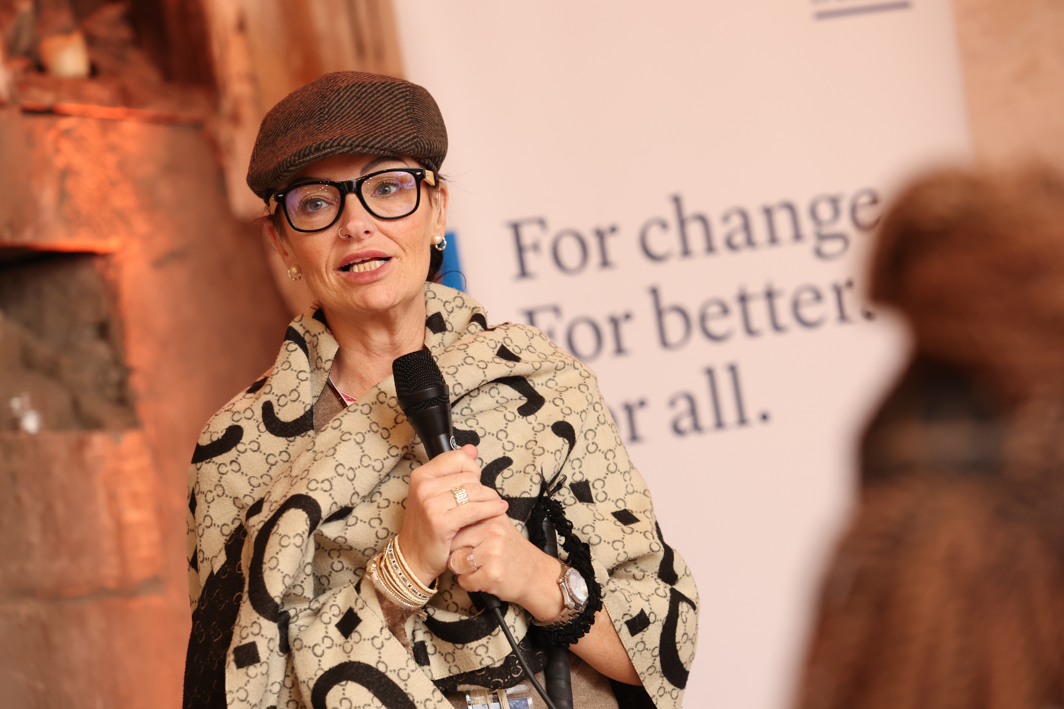 Image shows Julieanne Crothers holding a microphone and speaking. She wears a flat cap and has glasses.
