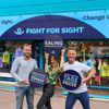Three people standing outside a Fight for Sight charity shop during the Ealing Fashion Drop, holding circular signs promoting new with tags fashion at charity shop prices and highlighting tonnes of clothing saved from landfill.